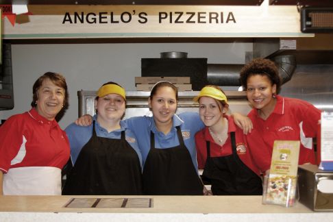 The female staff of Angelo's Pizzeria stand in a line behind the counter, smiling at the camera. The first woman has short brown hair and wears a red polo and white apron. The second, Brianna, is wearing a yellow hat, blue polo, and black apron. The third has her hair pulled back and is wearing a blue polo and black apron. The fourth is wearing a yellow hat, red polo, and black apron. And the fifth and final woman has curly brown hair and is wearing a red polo and white apron.