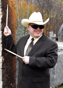 Roger is smiling, standing outside by a rock wall and holding drumsticks. He is wearing a white cowboy hat and dark glasses, a black suit jacket, tie, and white button-up shirt.