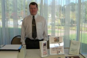 Roger is at a trade show, standing behind a table set with a printer, a small stack of brochures, a sign for Seeing Hands, and another small stack of booklets. He is wearing a white buttonup shirt and dark tie with a gold tie clip.