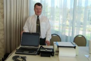 Roger is standing behind a white table set with a laptop and a few other pieces of adaptive electronics. He is wearing a white buttonup shirt and and dark colored tie with a gold clip. There is a window behind him overlooking a walkway and lots of greenery.