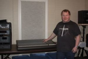 Roger stands in Studio C, near a Mackie 24-8 recording console. He is wearing a black shirt with a white cross on the front, and a silver wristwatch.