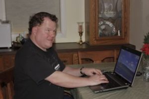 Roger sits at a table with an open laptop in front of him. He is wearing a black shirt and has a watch on his wrist.