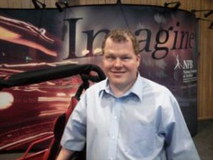 Roger stands in front of a black framed and red upholstered dune buggy. He is wearing a blue button-up shirt. There is a poster behind him depicting moving light on one side, with the word "Imagine" in large font across the top, and the NFB's logo on the left side.