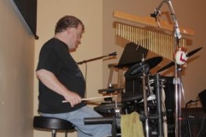 Roger sits behind an electronic drum set in a tan colored room. He is wearing a black shirt and jeans.